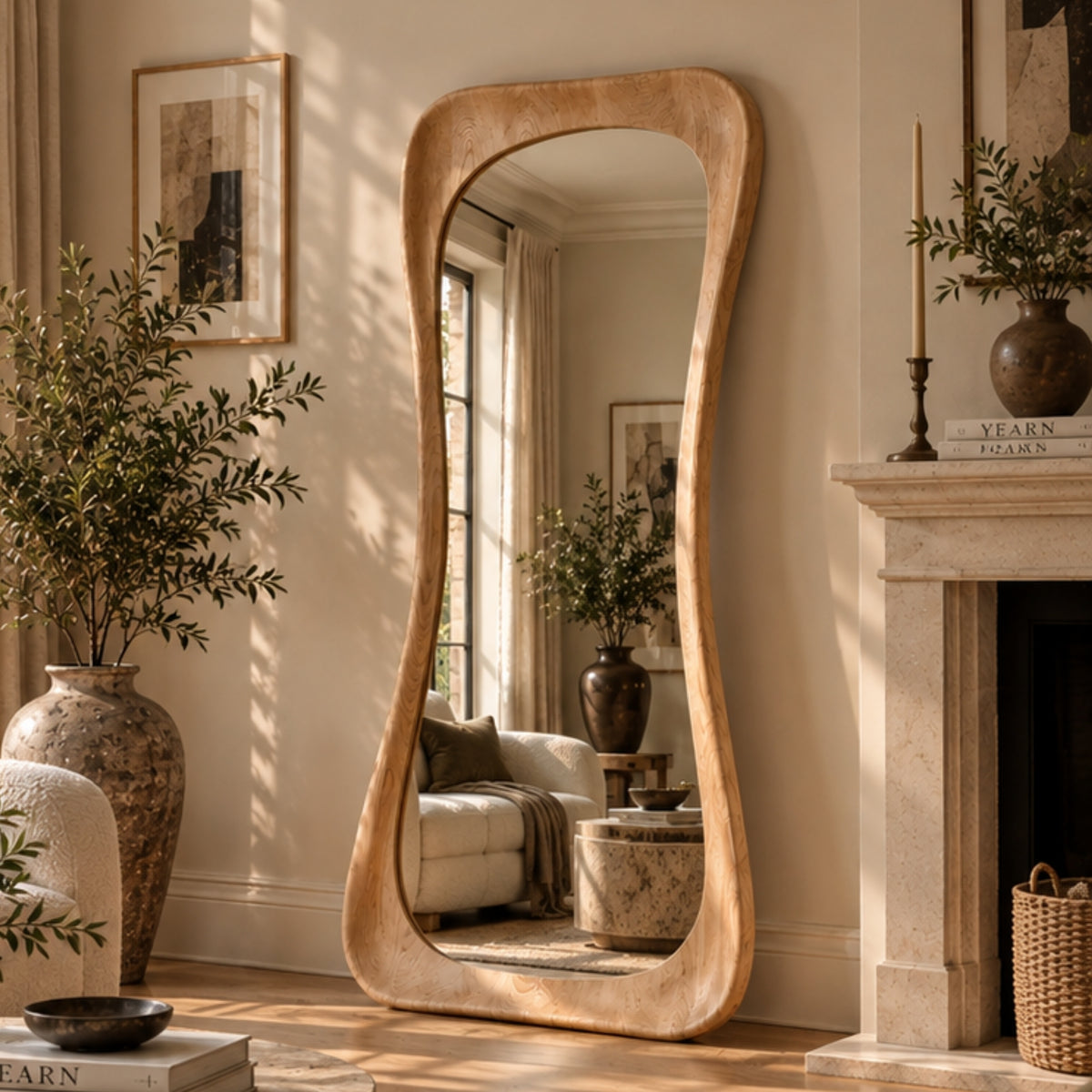 A tall, wavy-top floor mirror with a thick natural wood frame stands in a sunlit living room beside a fireplace and large potted plants.