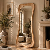 A tall, wavy-top floor mirror with a thick natural wood frame stands in a sunlit living room beside a fireplace and large potted plants.