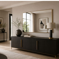 A modern black sideboard or credenza with slim gold-tone handles and feet, styled in a warm neutral living room with a large gold-framed mirror, table lamp, and decorative vases.