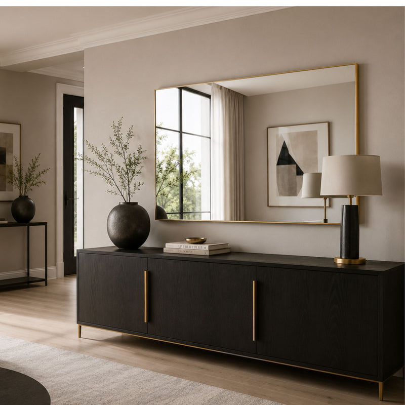 A modern black sideboard or credenza with slim gold-tone handles and feet, styled in a warm neutral living room with a large gold-framed mirror, table lamp, and decorative vases.