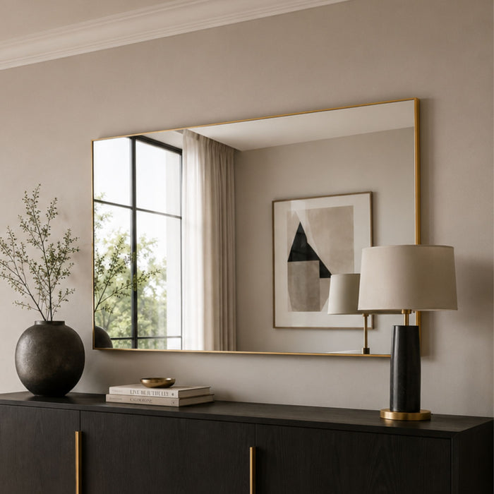 A large rectangular wall mirror with a thin gold frame hangs above a dark wood console table in a styled interior. The mirror reflects a window, curtain, framed abstract artwork, and a table lamp.