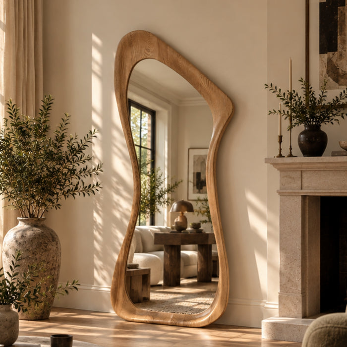 An organic-shaped full-length floor mirror with a natural oak wood frame stands in a sunlit room next to a fireplace and large potted plants.