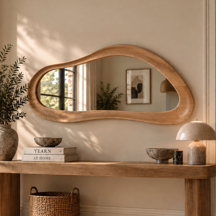 An organic-shaped wooden wall mirror with a thick natural oak frame, mounted on a beige wall above a matching wooden console table decorated with books, a vase, bowls, and a mushroom lamp.