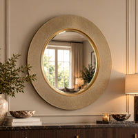 a round wall mirror with a textured, sunburst-style gold frame hangs on a beige panelled wall above a console table decorated with books, a vase, and a lit lamp.