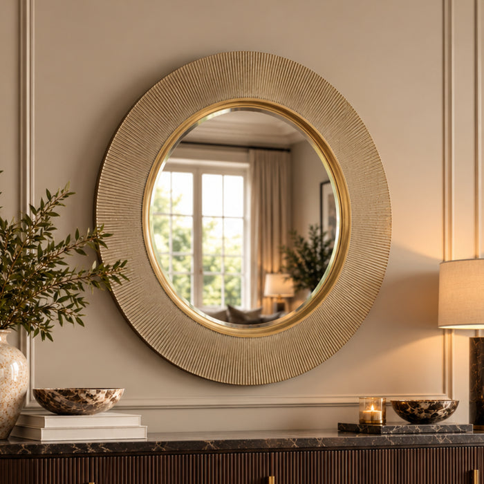 a round wall mirror with a textured, sunburst-style gold frame hangs on a beige panelled wall above a console table decorated with books, a vase, and a lit lamp.