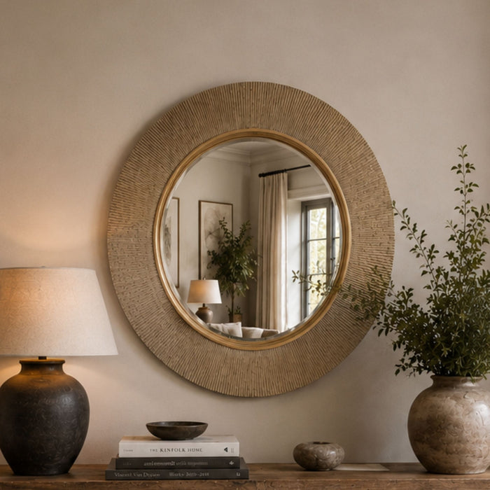 Round wall mirror with a textured beige woven frame and a gold-toned inner border, displayed on a console table in a styled room setting with a lamp, books, and a potted plant.