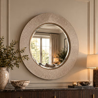 A large round wall mirror with a textured, sunburst-like metallic frame in a champagne gold finish, mounted on a beige paneled wall above a dark console table with decorative items.