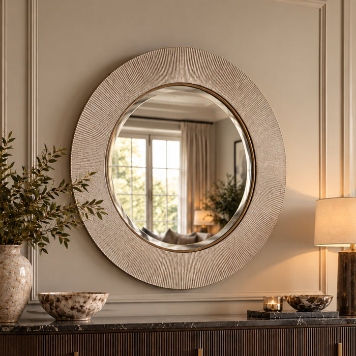 A large round wall mirror with a textured, sunburst-like metallic frame in a champagne gold finish, mounted on a beige paneled wall above a dark console table with decorative items.