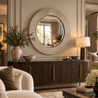A large round wall mirror with a textured, brushed silver frame hangs above a dark wood console table in a styled living room setting.