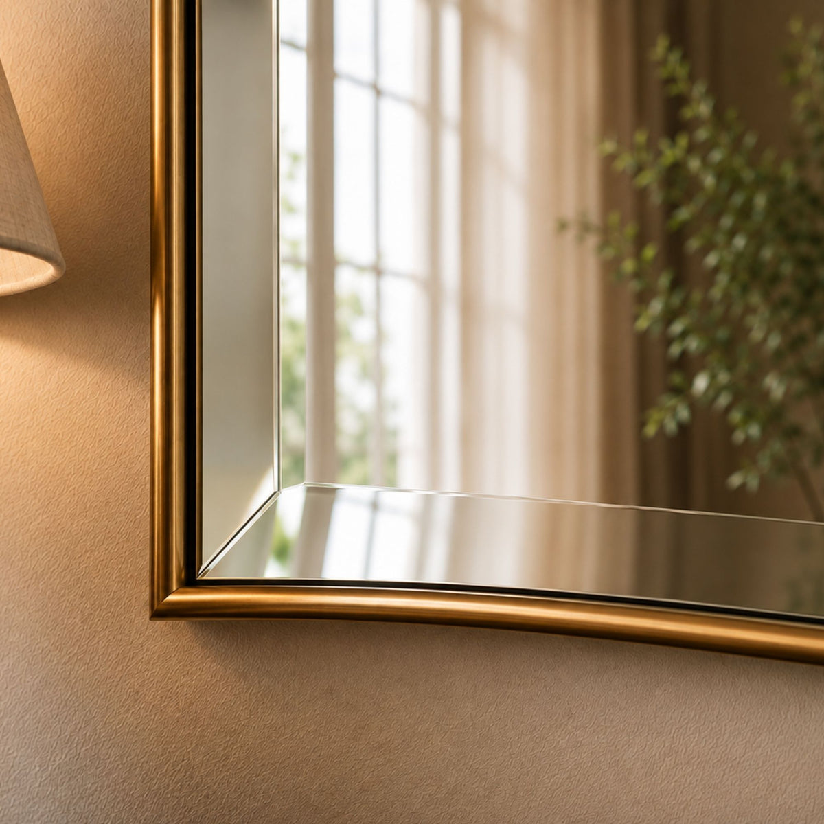 A close-up of a wall-mounted rectangular mirror with a brushed gold frame, reflecting a softly lit room with curtains, a plant, and a lamp.