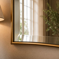 A close-up of a wall-mounted rectangular mirror with a brushed gold frame, reflecting a softly lit room with curtains, a plant, and a lamp.