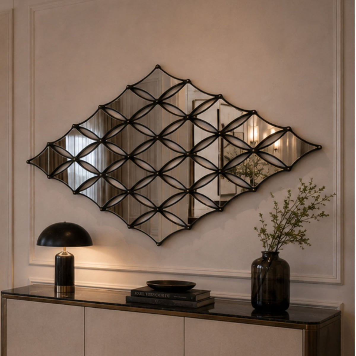 A large diamond-shaped wall mirror with an intricate black metal geometric frame hangs on a beige wall above a console table styled with a lamp, books, and a vase.