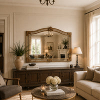 A large ornate gold-framed mirror hangs on a cream wall above a dark wood console in a styled living room. The room features a beige sofa, a marble-top coffee table with white flowers, a table lamp, and a vase of greenery.