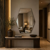 A modern hexagonal wall mirror with a thin dark frame hangs above a console table in a styled interior. The mirror reflects a softly lit living room with a sofa and lamp.