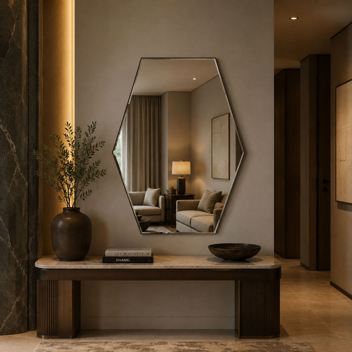 A modern hexagonal wall mirror with a thin dark frame hangs above a console table in a styled interior. The mirror reflects a softly lit living room with a sofa and lamp.