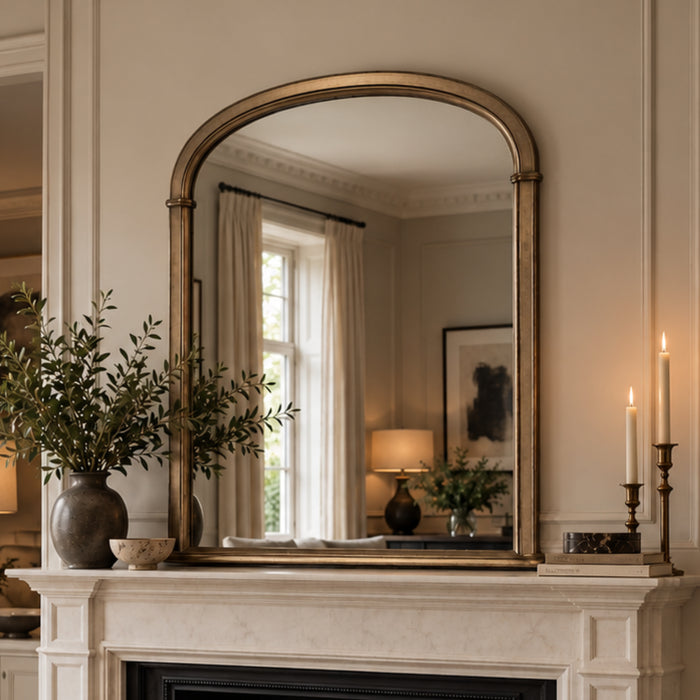 An arched wall mirror with an antique gold frame hanging above a white marble fireplace mantel in a warm, elegant room.