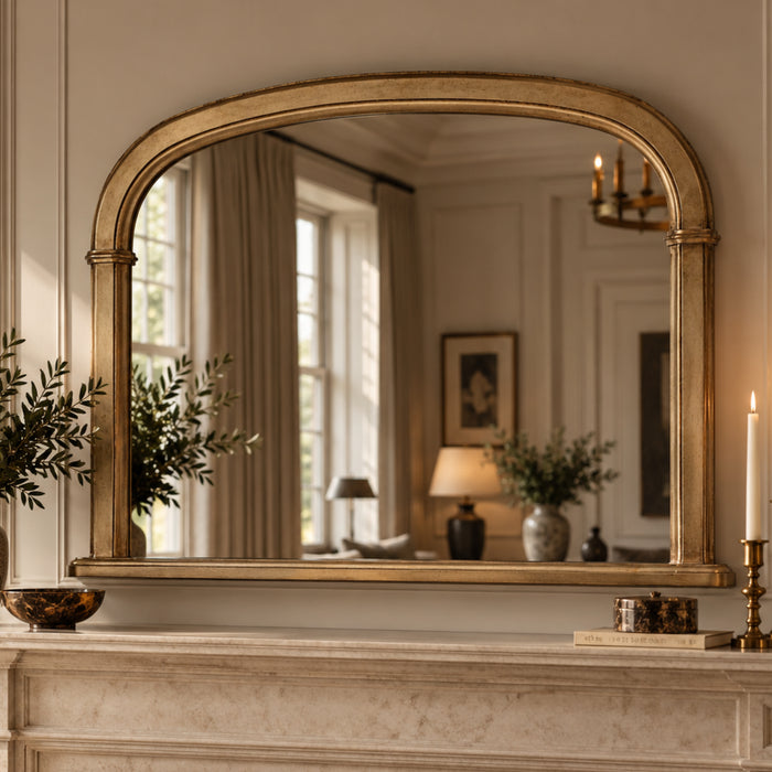 An arched gold-framed mirror with a distressed antique finish, mounted on a cream marble mantelpiece in a classic, elegant room. The mirror reflects a softly lit interior with beige curtains, a table lamp, and framed wall art.