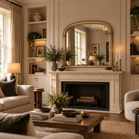 A beautifully styled living room featuring a large arched gold-framed mirror mounted above a classic stone fireplace. The room is decorated with warm neutral tones, including beige walls, cream sofas, a wooden coffee table, and built-in shelving with books and greenery.