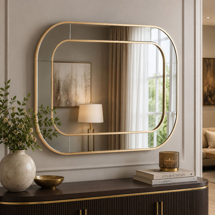 A large rectangular wall mirror with rounded corners and a gold frame, mounted on a beige wall above a dark wood console table. The mirror reflects a softly lit interior with curtains, a lamp, and artwork.