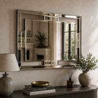 A large rectangular wall mirror with a polished silver frame hangs on a beige wall above a dark wooden console table. The scene includes a table lamp, books, a small diffuser, and potted plants, with the mirror reflecting an interior fireplace and window view.