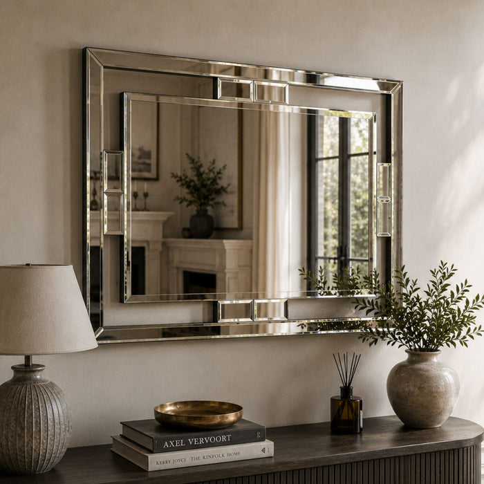 A large rectangular wall mirror with a polished silver frame hangs on a beige wall above a dark wooden console table. The scene includes a table lamp, books, a small diffuser, and potted plants, with the mirror reflecting an interior fireplace and window view.