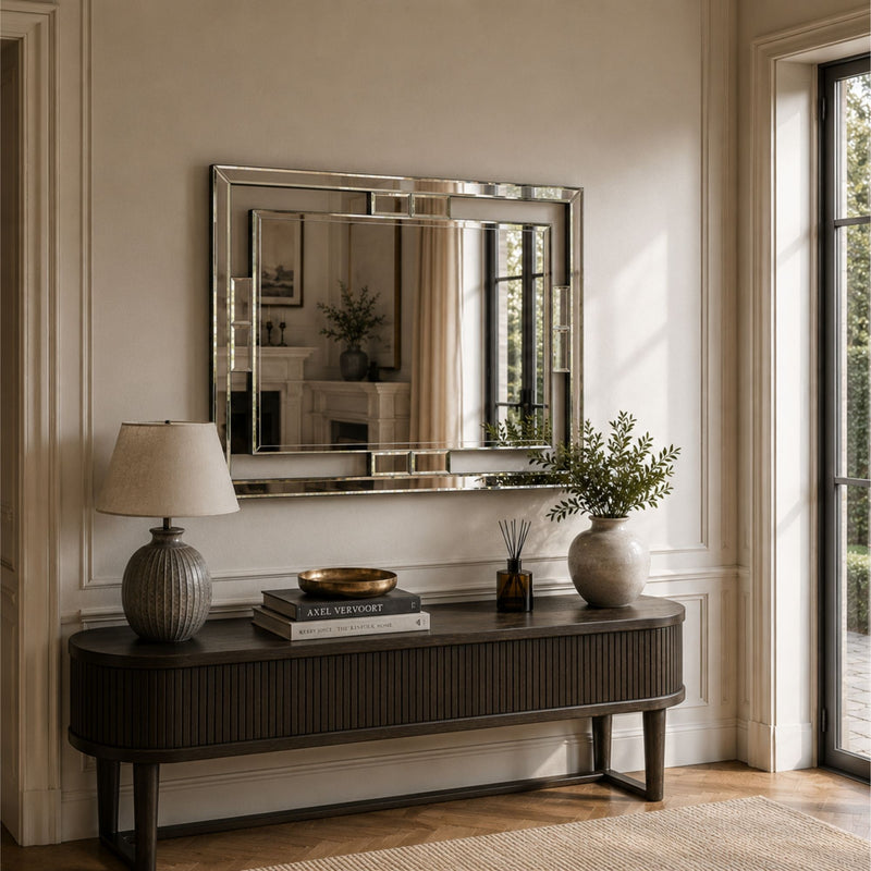 A large rectangular wall mirror with a polished silver frame hangs above a dark wood console table in a bright, elegant room with beige walls, a table lamp, books, and a vase.