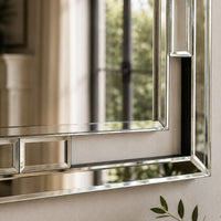 A close-up of a rectangular wall mirror with a polished silver-toned frame, reflecting a softly blurred interior and window view.