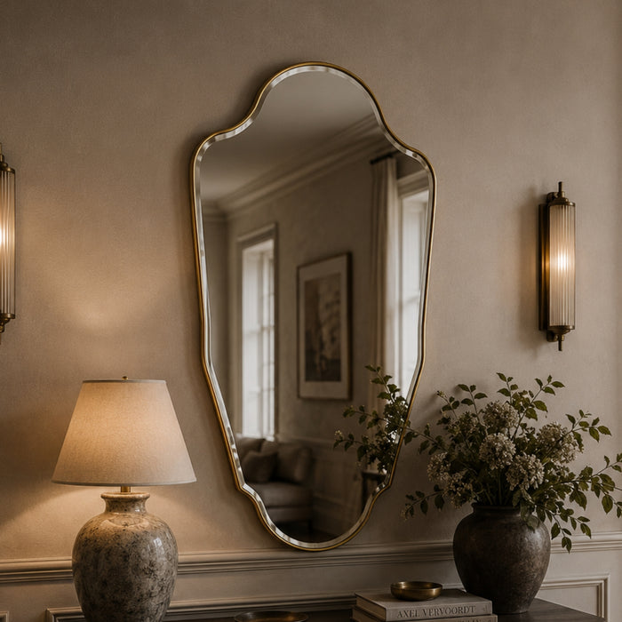 An ornate asymmetrical wall mirror with a gold-toned frame hangs on a beige wall above a console table. The mirror reflects a softly lit interior, while the room is styled with a lit table lamp, wall sconces, a vase of flowers, and books.