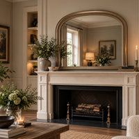 A large arched mirror with a gold-toned frame hangs above a cream fireplace mantel in a styled living room. The scene includes a black fireplace opening with logs, potted greenery on the mantel and table, framed wall art, candles, and warm ambient lighting.