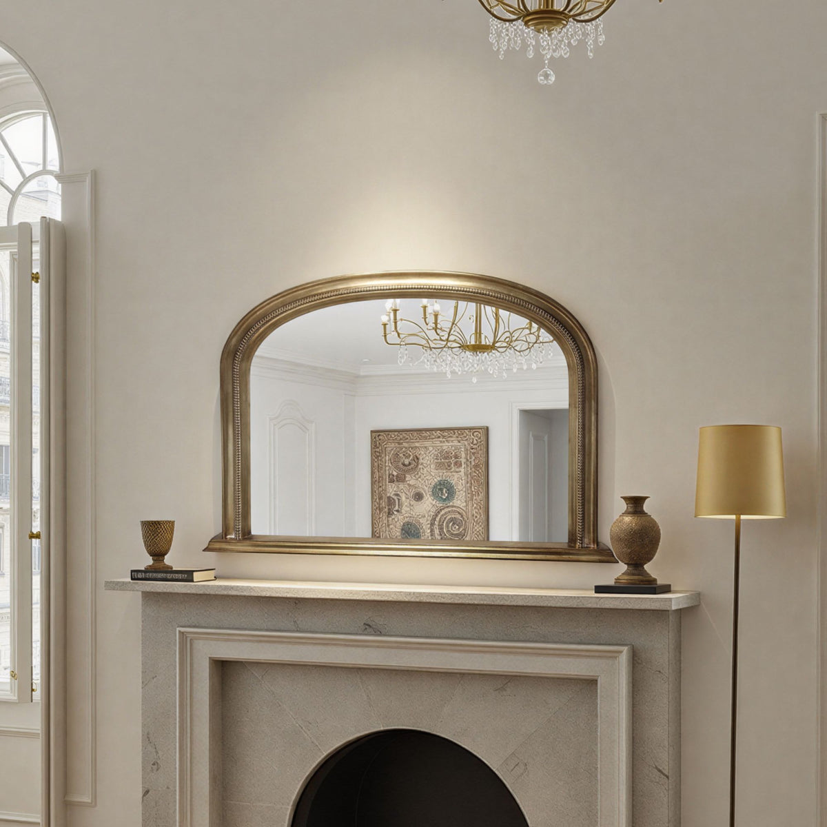 Decorative antique mirror above a fireplace with a chandelier reflected in it, in a room with a lamp and framed picture.