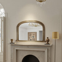 Decorative antique mirror above a fireplace with a chandelier reflected in it, in a room with a lamp and framed picture.