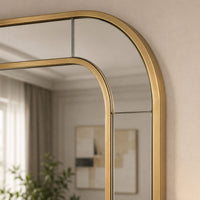 An arched wall mirror with a brushed gold metal frame, shown mounted on a beige wall with its reflection visible.