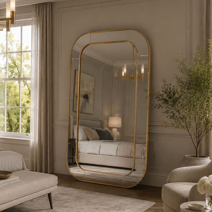 A large floor mirror with a gold frame and rounded top, styled in a cozy, elegant bedroom setting with beige walls, a bed, and soft natural light.