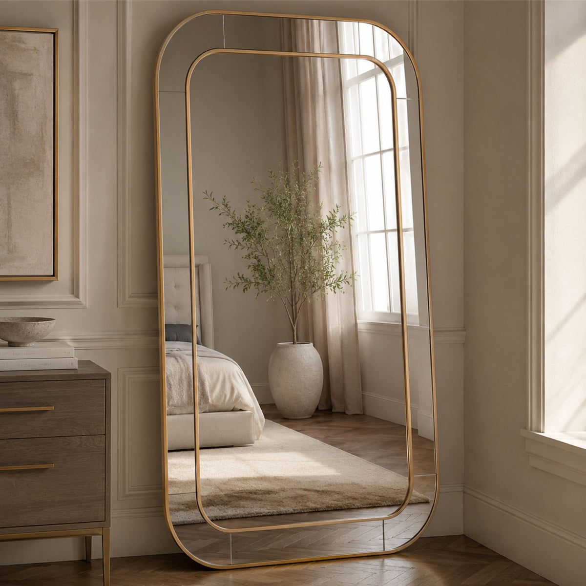 A tall floor mirror with a slim gold metal frame stands in a bedroom setting, reflecting a bed, curtains, and a potted plant.