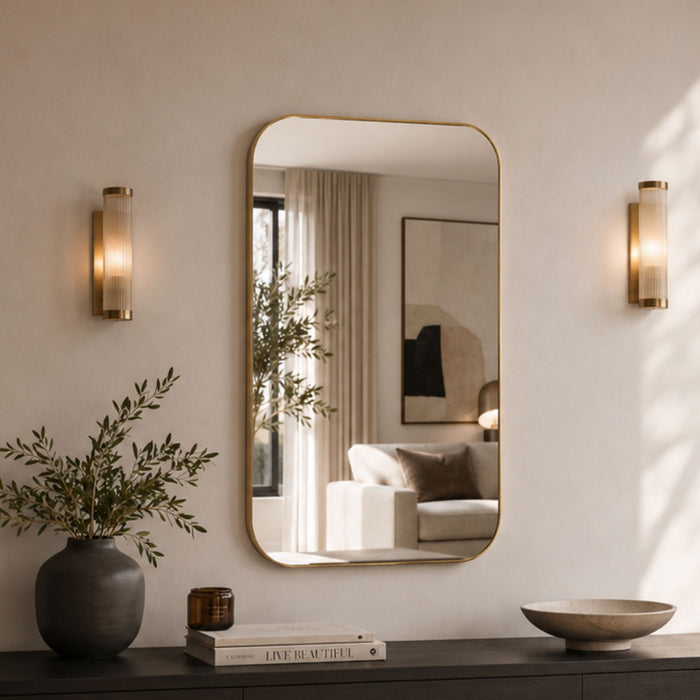 A large rounded rectangular wall mirror with a thin gold frame hangs on a beige wall, flanked by two matching gold wall sconces. The mirror reflects a softly lit living room interior, while the foreground shows a dark console table, a gray vase with greenery, a small amber candle, a book, and a ceramic bowl.