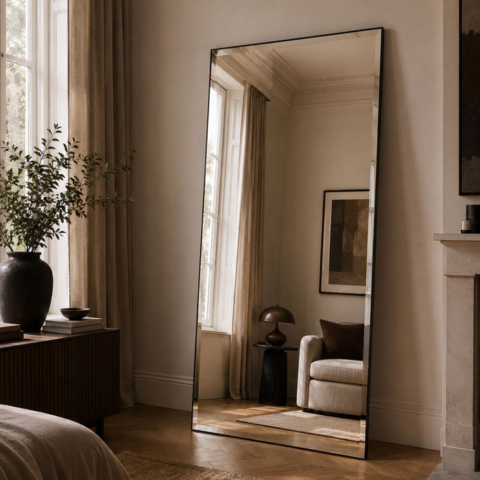 A tall floor mirror with a thin black frame stands in a softly lit bedroom or living space, reflecting a chair, lamp, and window. The room has beige walls, wood flooring, a dark vase with greenery on a sideboard, and a fireplace mantel.
