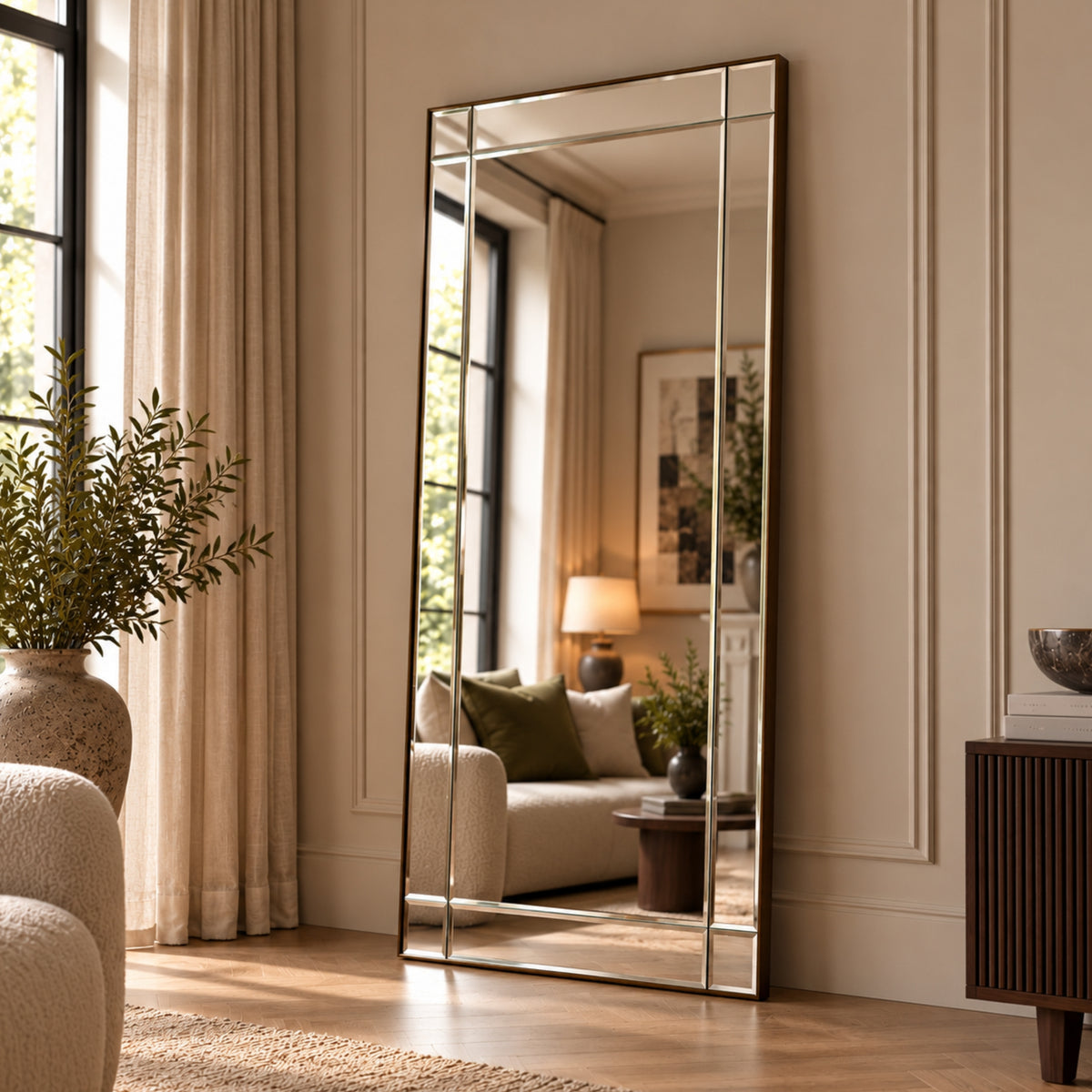 A tall full-length floor mirror with a thin metallic frame leans against a cream paneled wall in a styled living room. The mirror reflects a sofa, lamp, and window, while the room itself features beige curtains, a potted plant, wood flooring, and a dark wood sideboard.