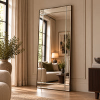 A tall full-length floor mirror with a thin metallic frame leans against a cream paneled wall in a styled living room. The mirror reflects a sofa, lamp, and window, while the room itself features beige curtains, a potted plant, wood flooring, and a dark wood sideboard.