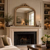 An ornate gold-framed arched mirror with a decorative crest top, mounted above a classic stone fireplace mantel in a beautifully styled living room.