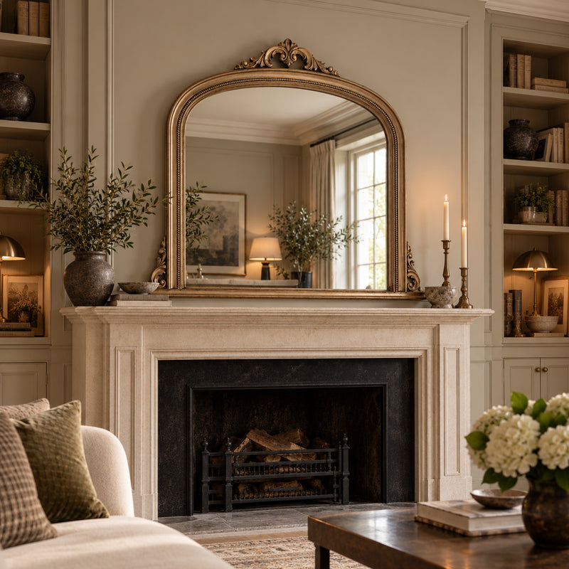 An ornate gold-framed arched mirror with a decorative crest top, mounted above a classic stone fireplace mantel in a beautifully styled living room.