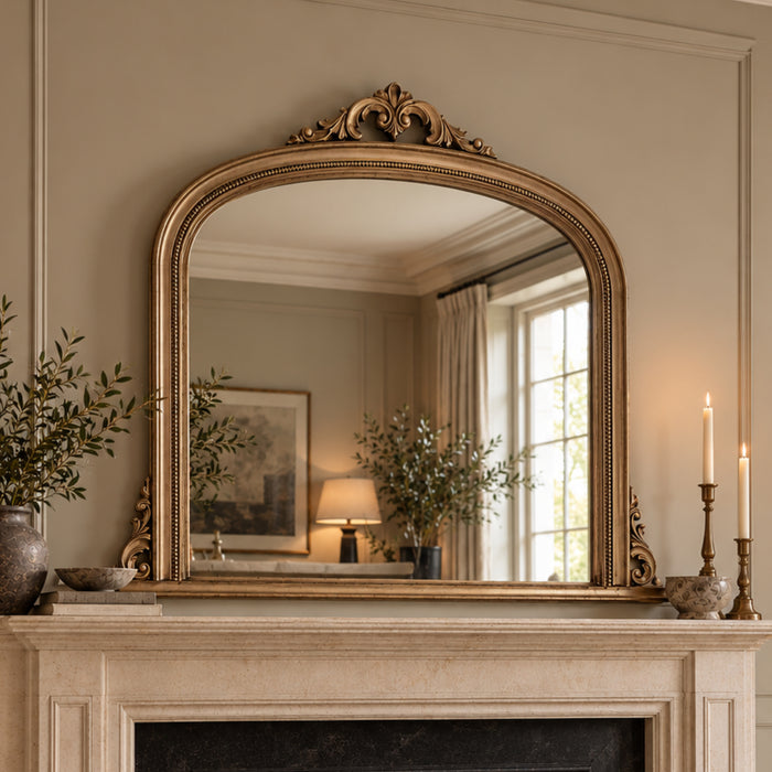 An ornate gold-framed arched mirror with a decorative crest top, mounted on a beige wall above a fireplace mantel. The mirror reflects a softly lit room with plants, a lamp, and a window, while candles sit on the mantel against the wall.