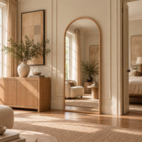 A tall arched floor mirror with a light wood frame stands in a sunlit, neutral-toned living room. The mirror reflects an armchair and side table, while the room features a light wood credenza, beige walls, framed art, and a woven rug.