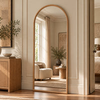 An arched full-length floor mirror with a light natural wood frame, standing in a beautifully styled bedroom doorway. The mirror reflects a cozy, sunlit room with a cream armchair, a side table, and a window, while the surrounding room features matching wood paneling, a wooden sideboard, and a textured beige rug.
