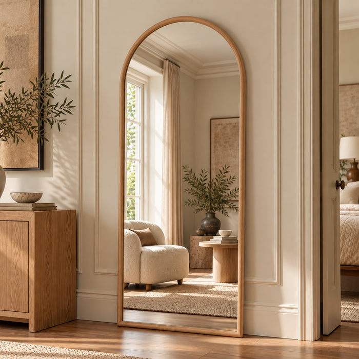An arched full-length floor mirror with a light natural wood frame, standing in a beautifully styled bedroom doorway. The mirror reflects a cozy, sunlit room with a cream armchair, a side table, and a window, while the surrounding room features matching wood paneling, a wooden sideboard, and a textured beige rug.