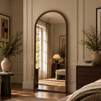 An arched full-length floor mirror with a warm wood frame, styled in a beautifully decorated bedroom with cream walls, a wooden dresser, and a large vase of greenery.