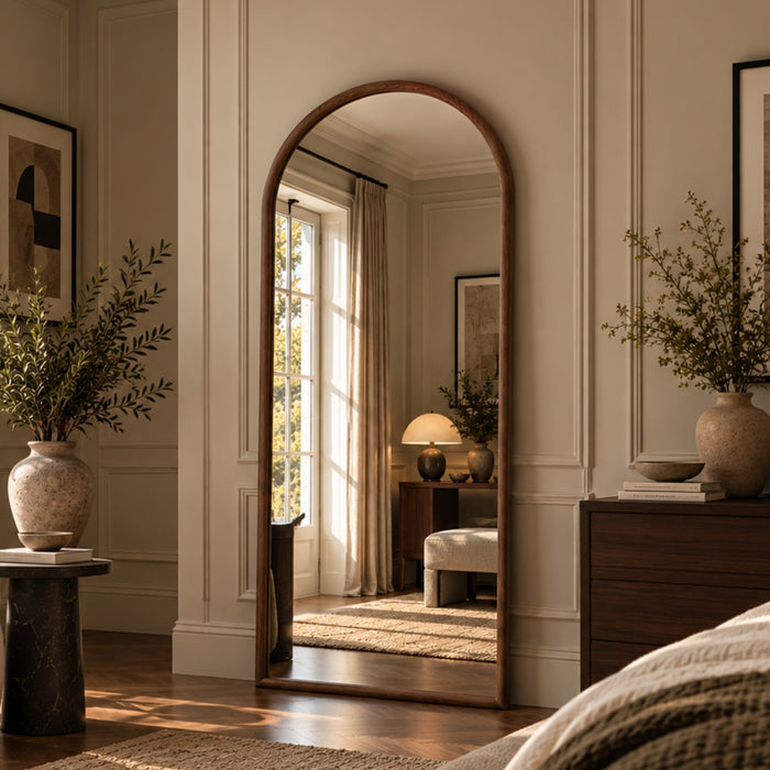 An arched full-length floor mirror with a warm wood frame, styled in a beautifully decorated bedroom with cream walls, a wooden dresser, and a large vase of greenery.