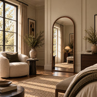 A styled interior room with warm beige walls, an arched floor mirror, a cream armchair, a dark wood dresser, and natural light streaming through tall windows.