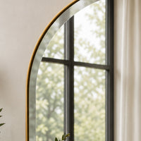 An arched wall mirror with a thin gold frame, reflecting a window view of green foliage, set against a neutral beige wall.