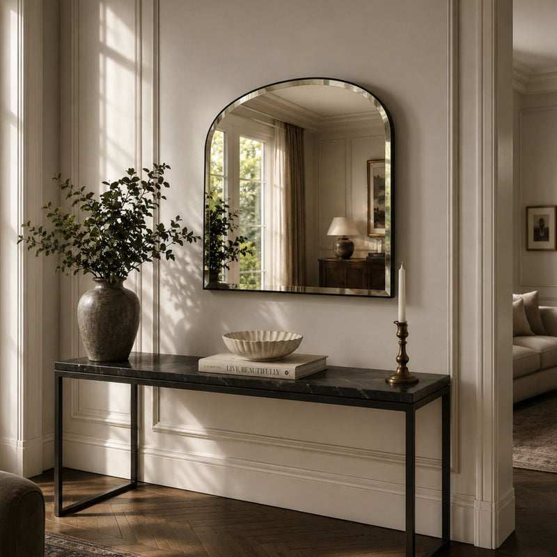 An arched wall mirror with a dark metal frame hangs above a console table in a styled interior room. The scene includes a vase with greenery, a bowl, candles, wood flooring, and a glimpse of a sofa in the background.