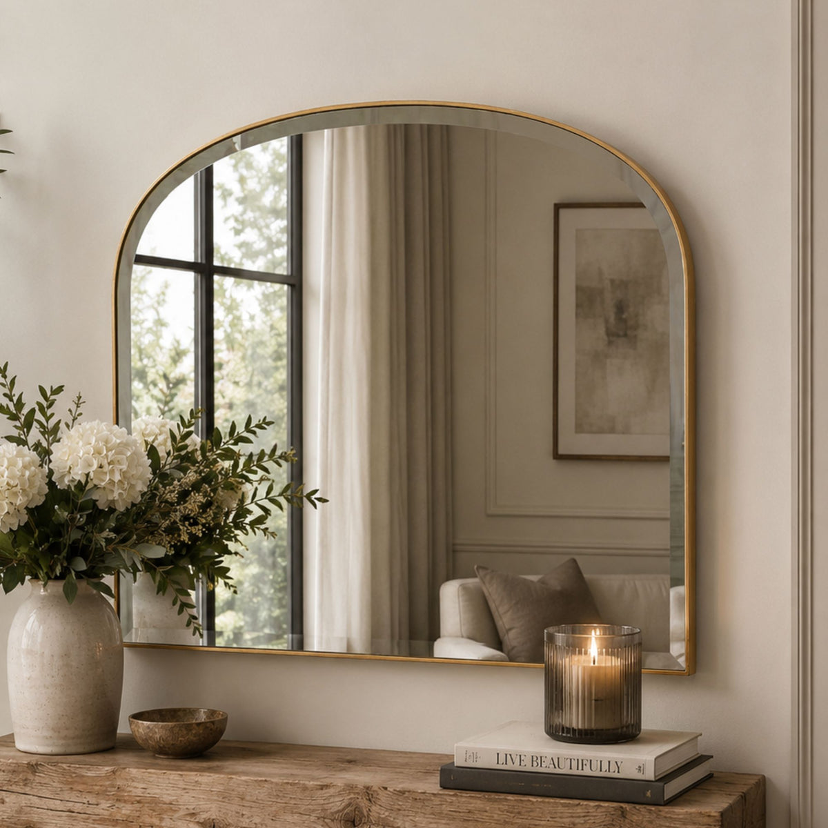 An arched wall mirror with a thin gold frame hangs above a rustic wooden console table. The mirror reflects a softly lit interior with curtains, a framed print, and a chair, while the room itself is styled with a large white flower arrangement in a textured vase and a lit candle on stacked books.
